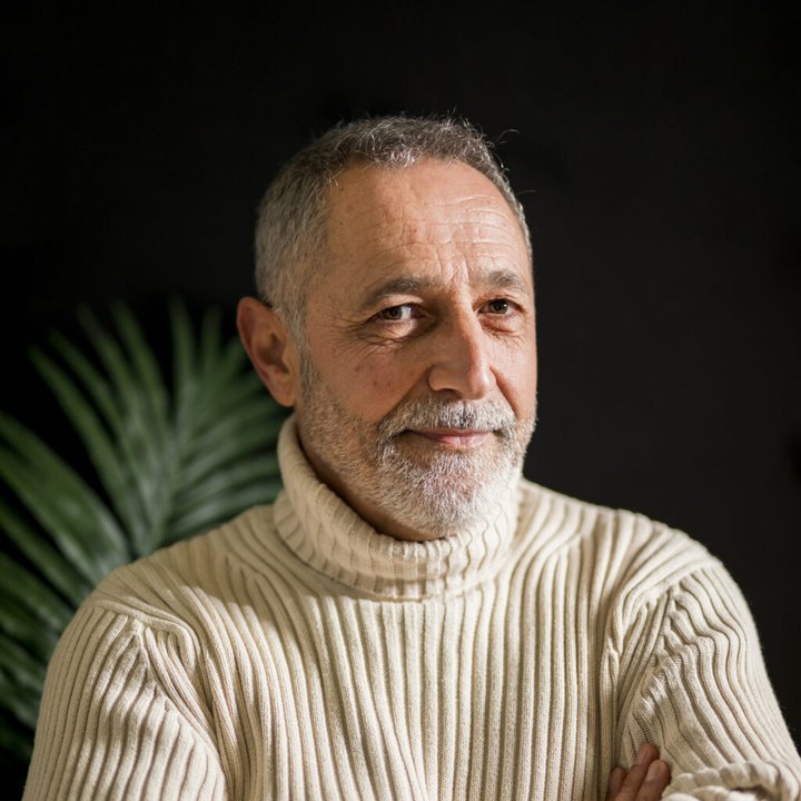 A calm man with a grey beard in a cream turtleneck beside a green plant